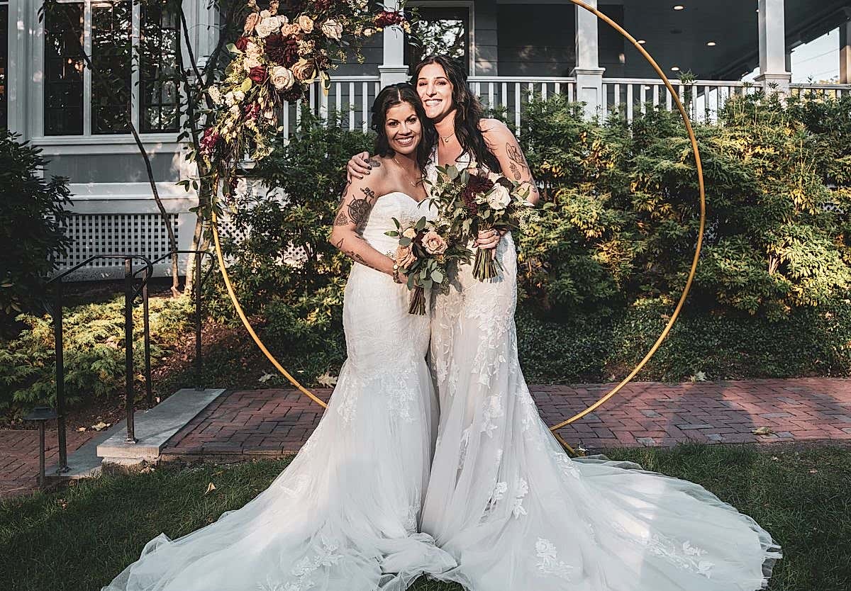 Two brides in white wedding gowns stand outdoors, smiling and holding bouquets in front of a floral arch with greenery in the background.