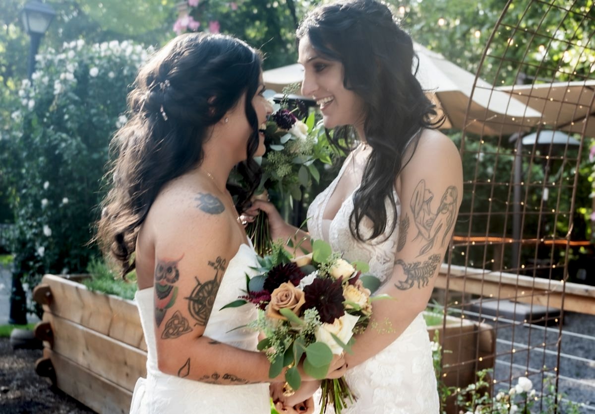 Two brides with tattoos stand close, smiling at each other, holding bouquets at an outdoor wedding surrounded by greenery and soft lights.