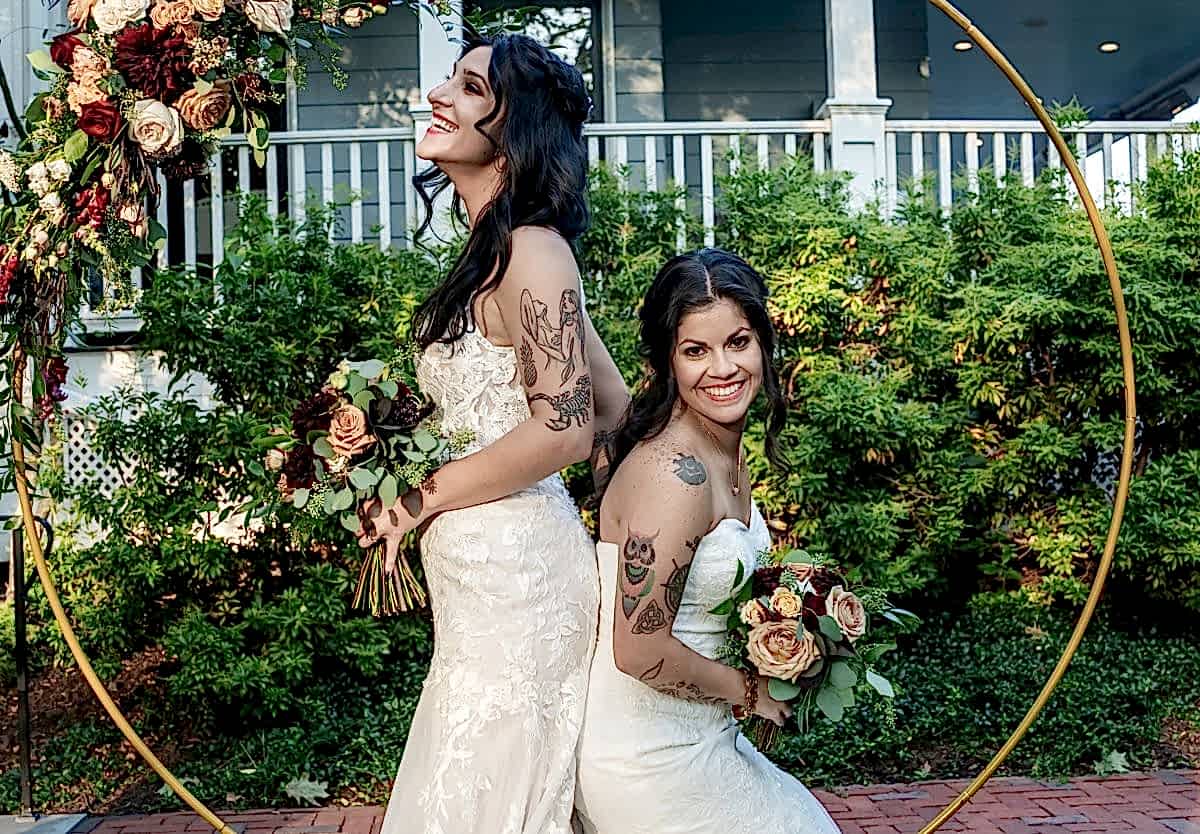 Two brides in white dresses sit back to back, holding bouquets, smiling in front of a floral arch and greenery outdoors.