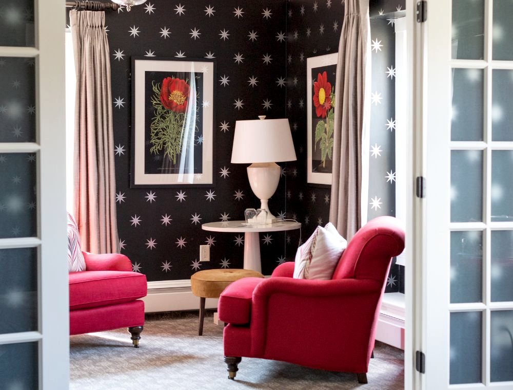 A cozy room with two red armchairs, a round table with a white lamp, star-patterned dark wallpaper, and flower artwork on the walls.