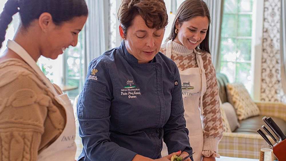 A chef in a blue jacket prepares food while two women in aprons watch and smile in a bright, cozy kitchen.