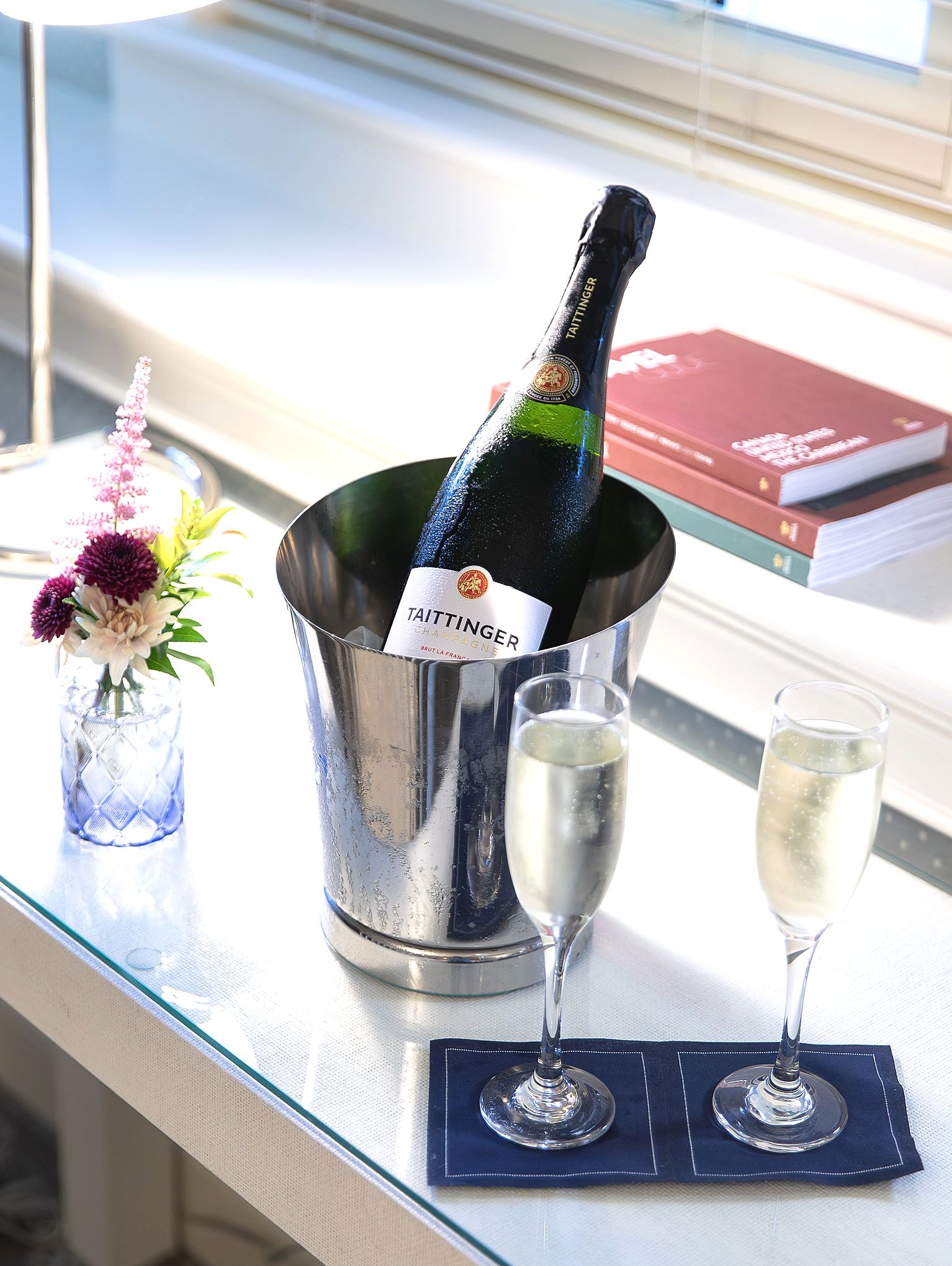 A bottle of Taittinger champagne in an ice bucket with two filled glasses, a small flower vase, and books on a white desk.