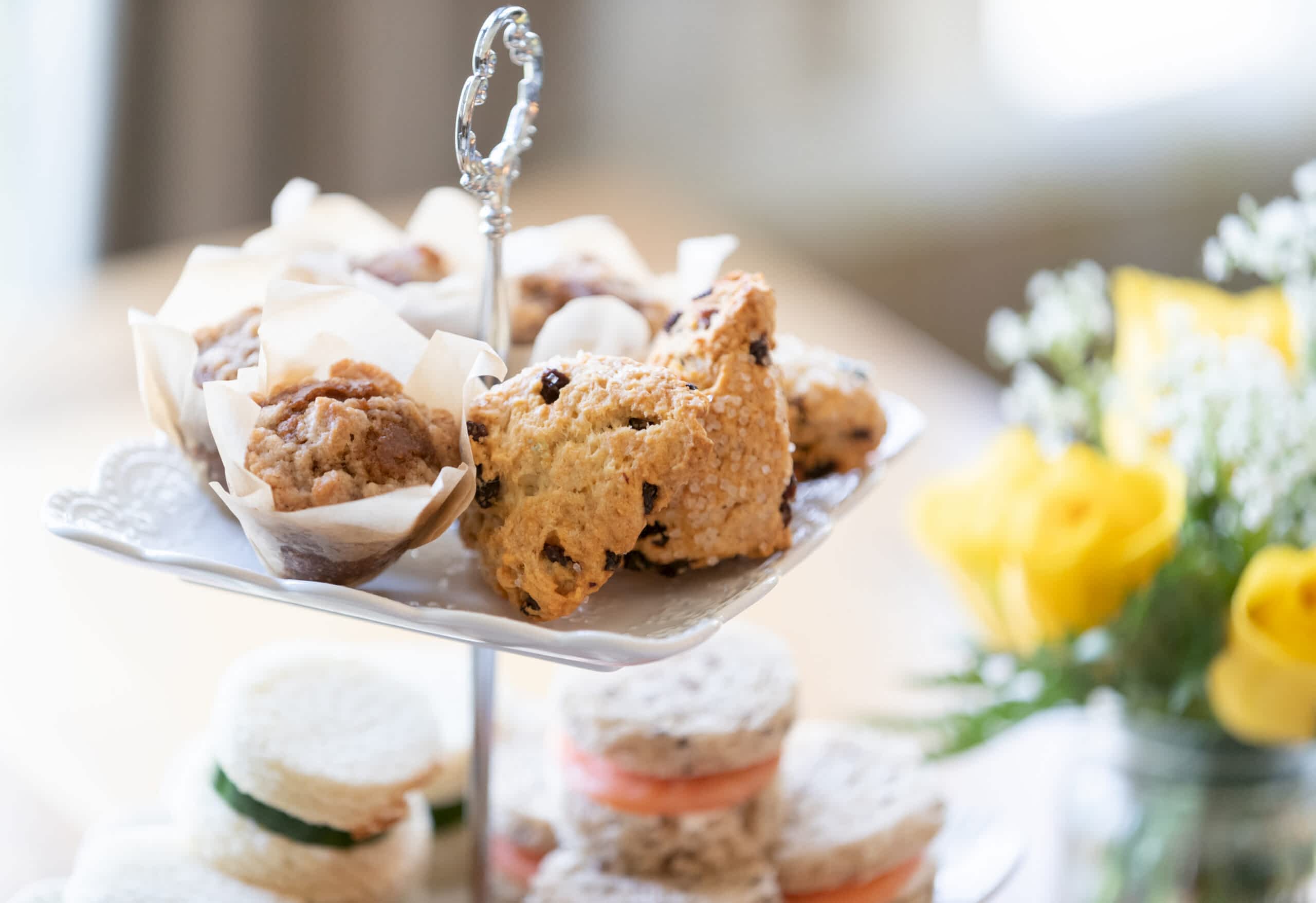 A tiered tray with muffins, scones, and finger sandwiches. Yellow flowers and more sandwiches are visible in the background.