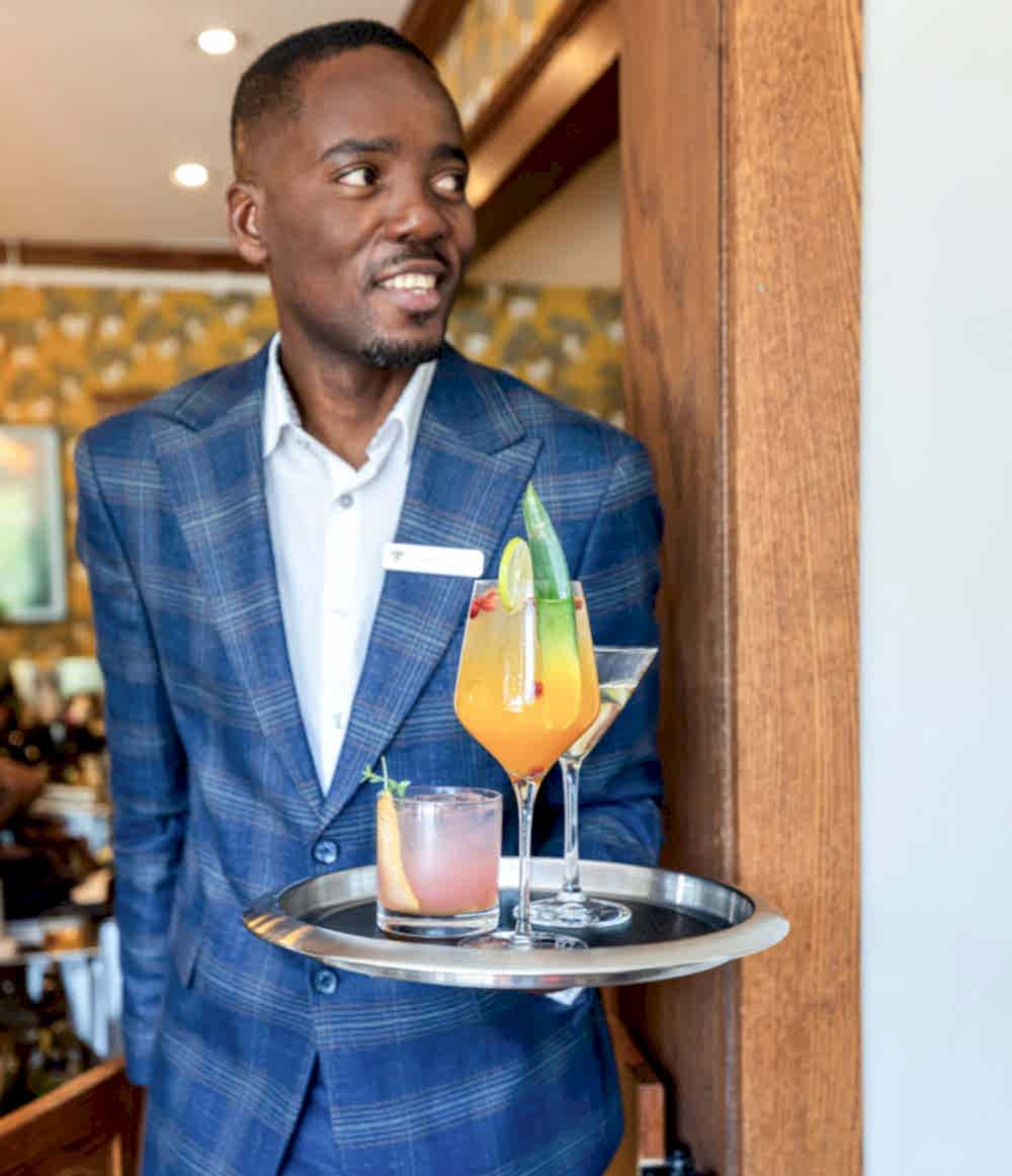 A smiling waiter in a blue plaid suit holds a tray with colorful cocktails, standing near a wooden door inside a restaurant.