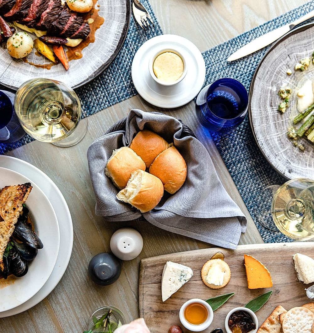 Bread rolls in a gray napkin surrounded by cheese, wine, seafood, steak, and vegetables on a dining table set for a meal.