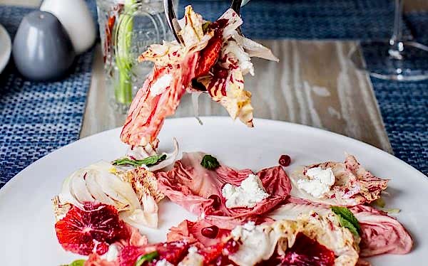 A hand serves salad with blood oranges, radicchio, goat cheese, fennel, and pomegranate seeds on a white plate.