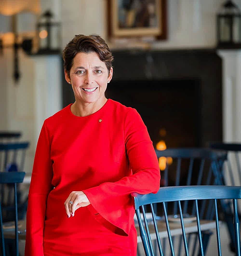 Woman in a bright red dress smiles while standing in a cozy room with blue chairs and a lit fireplace in the background.