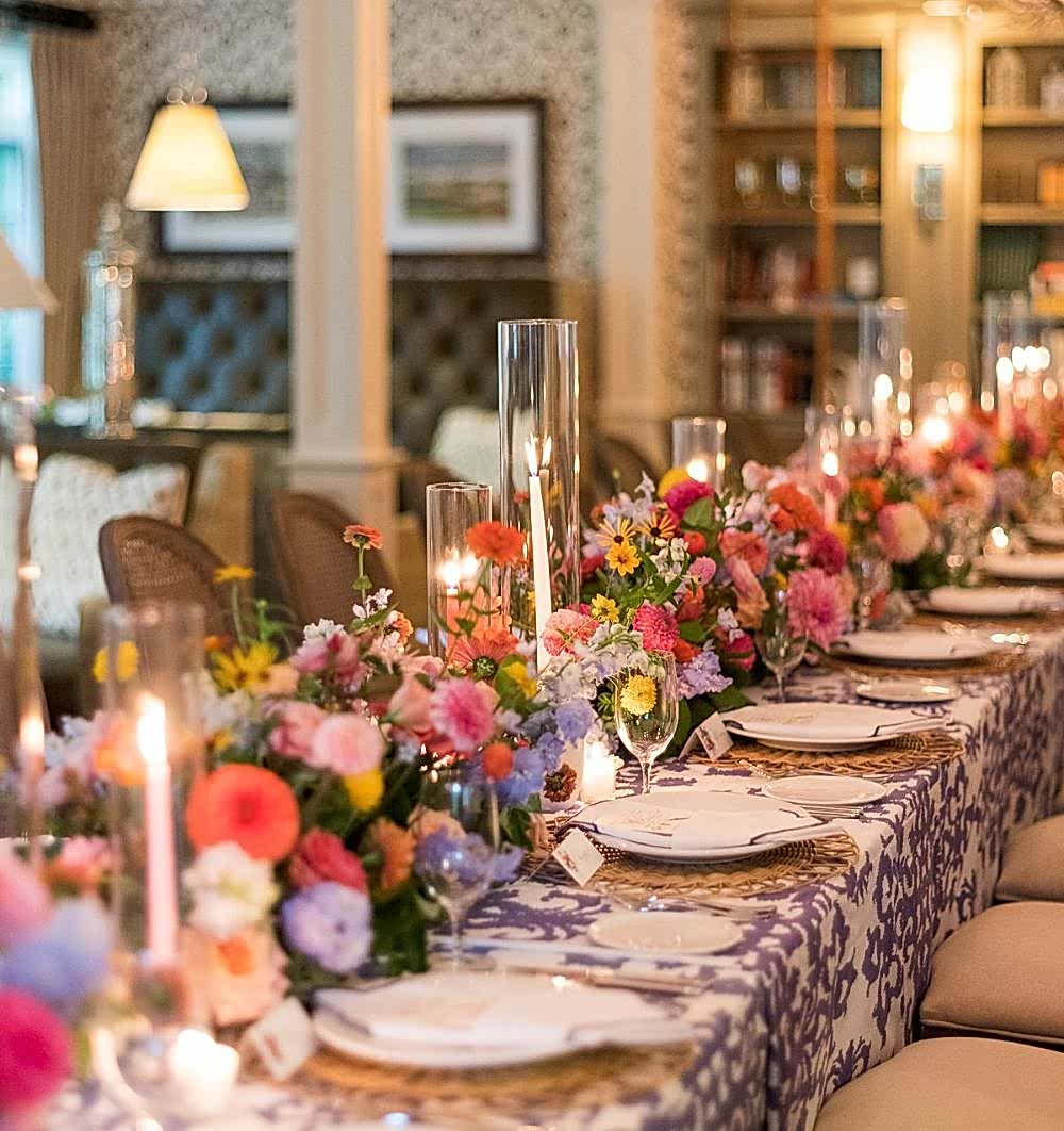A long dining table is set with floral arrangements, candles, plates, and glassware in an elegant, warmly lit room.