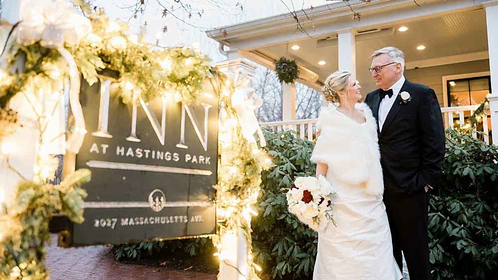 A bride and groom in formal wedding attire smile at each other outside the decorated Inn at Hastings Park on a winter day.