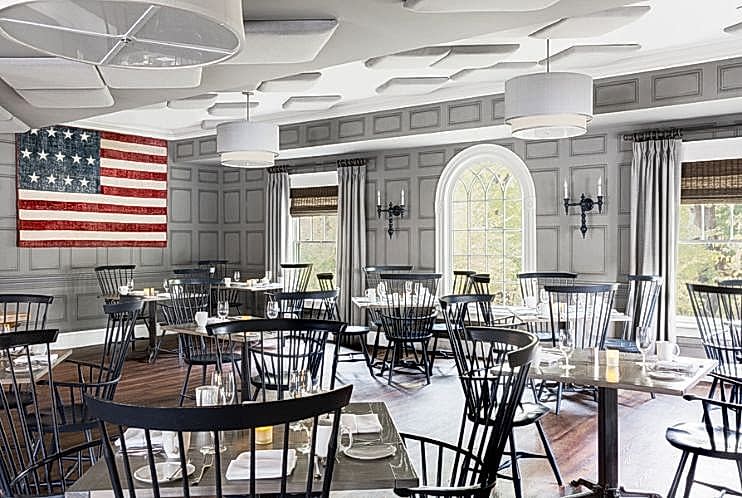 A bright, elegant dining room with black chairs, set tables, large windows, and an American flag on a gray paneled wall.