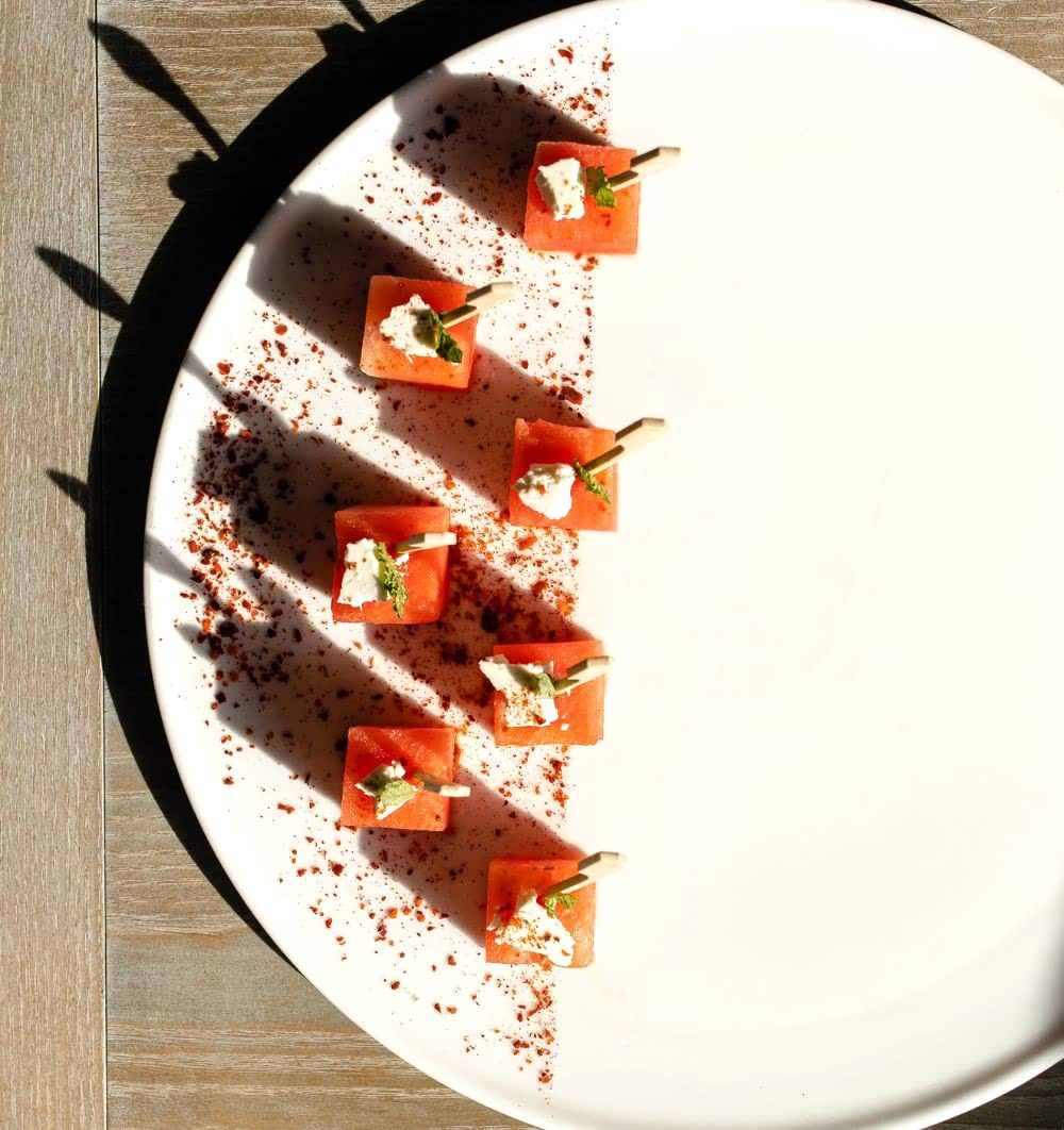Six cubes of watermelon with cheese and herbs on toothpicks, artfully arranged on a plate with red seasoning along one side, in bright sunlight.