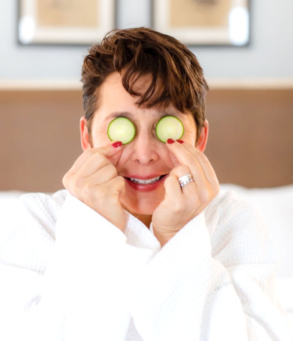 A woman in a white robe smiles while holding cucumber slices over her eyes, as if doing a spa treatment.