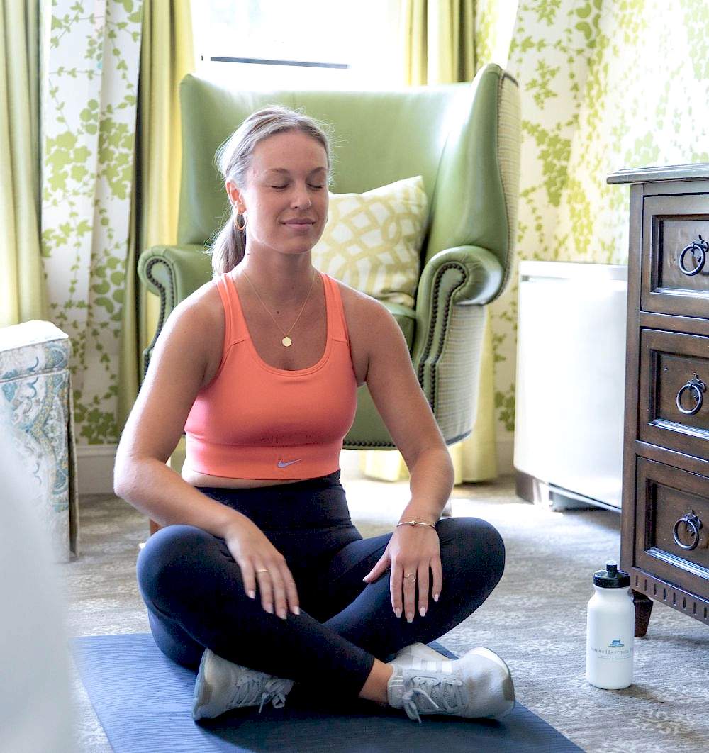 Woman in workout clothes sits cross-legged on a yoga mat indoors, eyes closed, appearing to meditate. A water bottle is on the floor nearby.